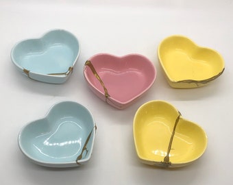 May include: Set of four heart-shaped bowls in light blue, pink, and yellow with gold accents. The bowls are made of ceramic and have a glossy finish.