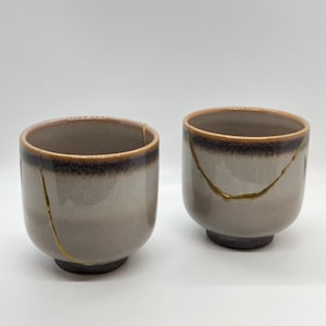 May include: Two ceramic teacups with a light gray glaze and a dark brown rim. The cups have gold cracks running through the glaze.