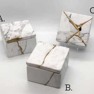 May include: Three white ceramic boxes with gold accents. The boxes have a marble-like pattern and are decorated with gold lines. The boxes are labeled A, B, and C.