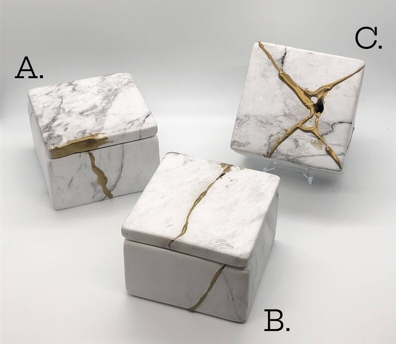 May include: Three white ceramic boxes with gold accents. The boxes have a marble-like pattern and are decorated with gold lines. The boxes are labeled A, B, and C.