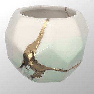 May include: White ceramic planter with a geometric shape and gold accents. The planter has a light green glaze and gold crackle lines.