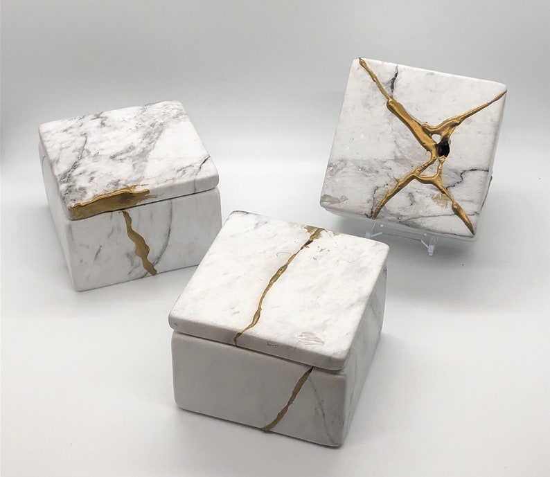 May include: Three white square boxes with gold accents. The boxes have a marble-like pattern and are decorated with gold lines. The boxes are different sizes and are arranged in a triangular shape.