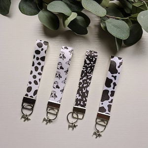 May include: Four black and white patterned keychains with cow-themed charms. The keychains feature different patterns, including cow spots, cow print, and a repeating cow illustration.