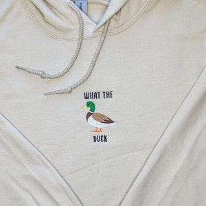 May include: A beige hoodie featuring the text "What the Duck" and an embroidered image of a green-headed duck.