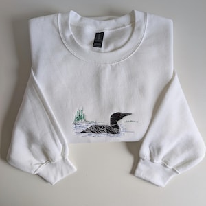 May include: White sweatshirt with a crew neck and long sleeves. The front features an embroidered design of a loon swimming in water, with green trees in the background. The loon has a black body, a black head, and a red eye.