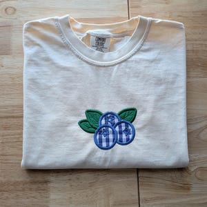 May include: A cream-colored short-sleeve t-shirt featuring an embroidered graphic of three blueberries with blue and white gingham patterns and green leaves. The inner neck label displays the text Comfort Colors, Heavyweight Garment Dye.