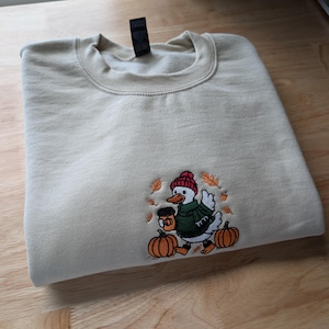 May include: A beige crewneck sweatshirt featuring an embroidered design of a white duck wearing a red knit hat and a green sweater. The duck holds a brown coffee cup, surrounded by orange pumpkins and falling autumn leaves in shades of gold.