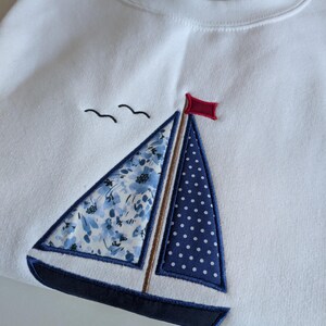 May include: White sweatshirt with a blue and white sailboat embroidered on the front. The sailboat has a red flag and a floral pattern on the sail.