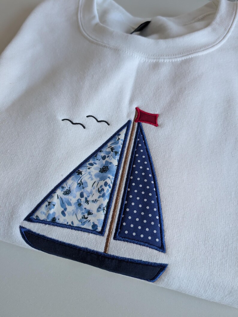 May include: White sweatshirt with a blue and white sailboat embroidered on the front. The sailboat has a red flag and a floral pattern on the sail.