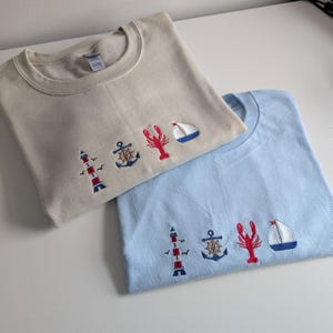 May include: Two t-shirts with nautical themed embroidery. The top shirt is beige with a lighthouse, anchor, lobster, and sailboat. The bottom shirt is light blue with a lighthouse, anchor, lobster, and sailboat.