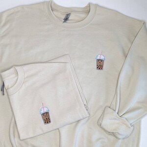 May include: Two beige sweatshirts with a brown bubble tea graphic embroidered on the front. The bubble tea has a pink straw and a smiling face.