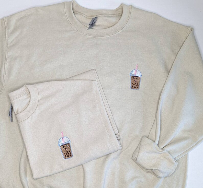 May include: Two beige sweatshirts with a brown bubble tea graphic embroidered on the front. The bubble tea has a pink straw and a smiling face.