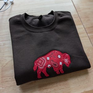 May include: A dark brown crewneck sweatshirt featuring a large, embroidered patch of a buffalo in profile. The buffalo is filled with a vibrant red paisley bandana pattern that includes white floral and teardrop motifs across its entire body.