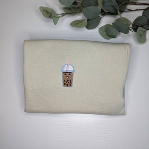 May include: A beige sweatshirt with a brown and white embroidered boba tea cup with a pink straw and a smiling face.