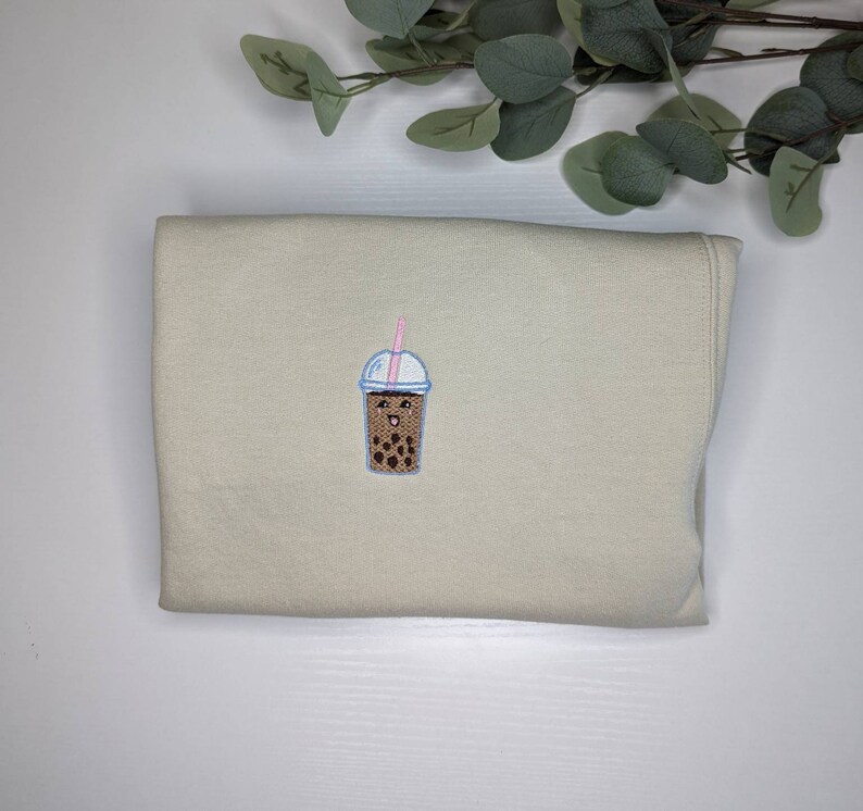 May include: A beige sweatshirt with a brown and white embroidered boba tea cup with a pink straw and a smiling face.