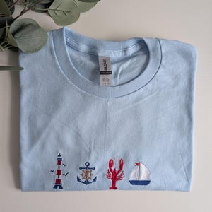 May include: Light blue t-shirt with embroidered nautical themed images including a lighthouse, anchor, lobster, and sailboat.