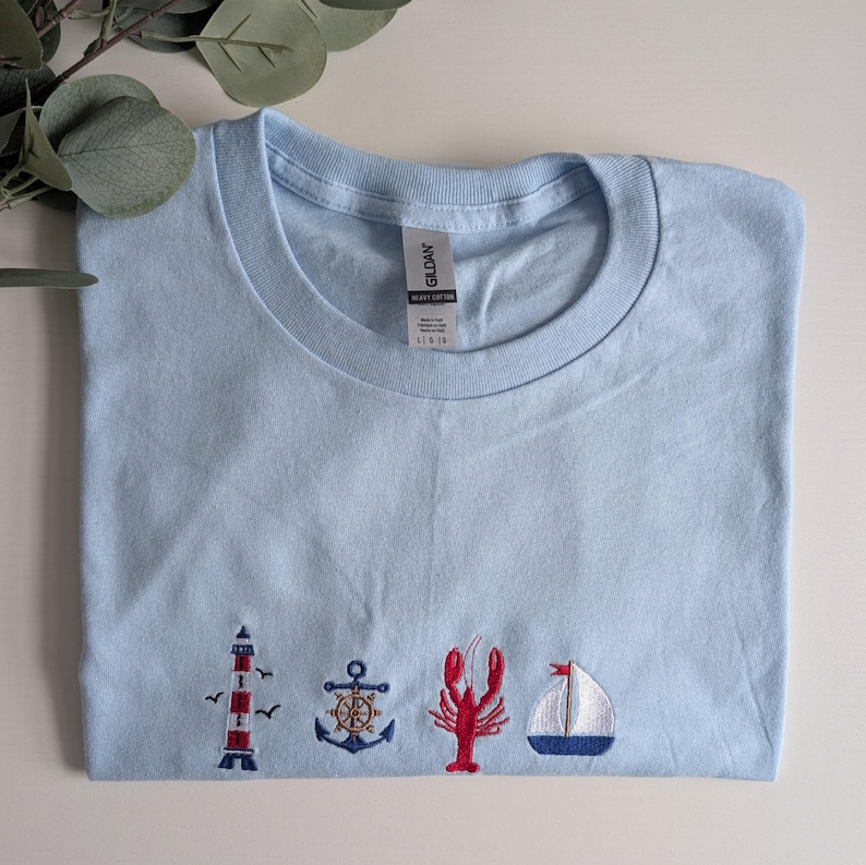 May include: Light blue t-shirt with embroidered nautical themed images including a lighthouse, anchor, lobster, and sailboat.