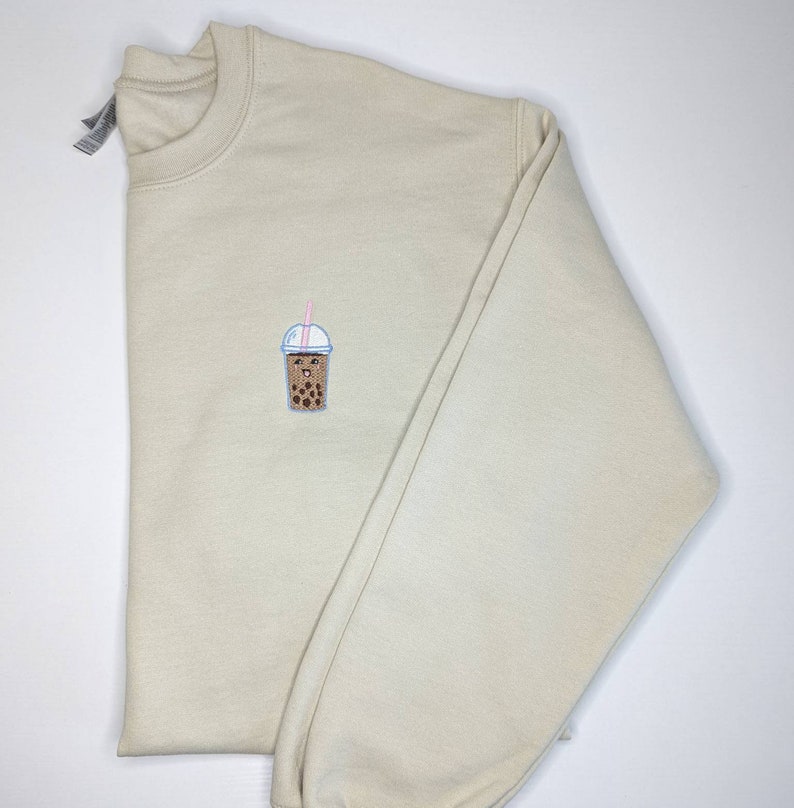 May include: A beige sweatshirt with a small embroidered image of a brown bubble tea cup with a pink straw and a smiling face.