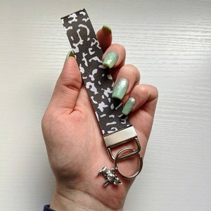 May include: A brown and white leopard print key chain with a silver key ring and a silver charm of a horse. The key chain is held in a hand with light green nail polish.