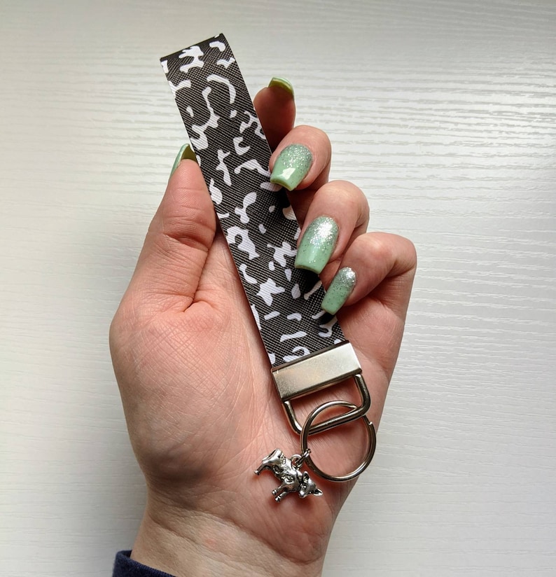 May include: A brown and white leopard print key chain with a silver key ring and a silver charm of a horse. The key chain is held in a hand with light green nail polish.