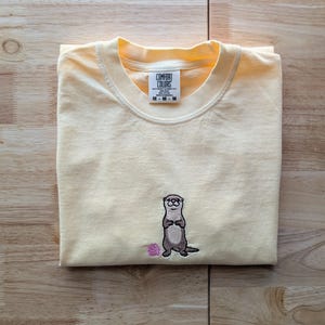 May include: A folded, light yellow t-shirt with a small embroidered otter design. The otter is brown and gray, holding a pink seashell. The shirt has a Comfort Colors tag.
