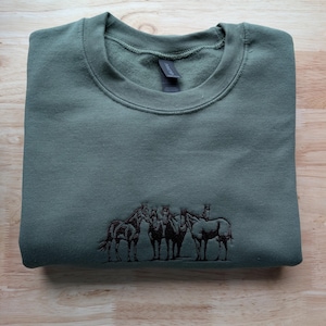 Puede incluir: Una sudadera de cuello redondo en color verde oliva con un gráfico bordado en marrón oscuro de cuatro caballos de pie en fila. La prenda cuenta con un cuello acanalado y una etiqueta de marca visible en el interior del escote.