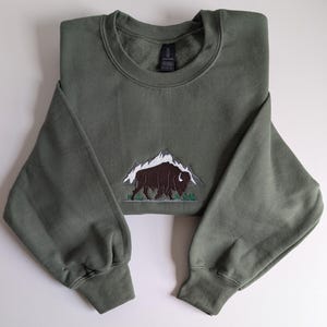 Könnte beinhalten: Olivgrünes Sweatshirt mit einem gestickten Design eines Bisons vor einer Bergkette. Der Bison ist braun, die Berge sind weiß. Das Sweatshirt hat einen Rundhalsausschnitt und lange Ärmel.