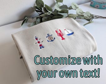 May include: A folded beige sweatshirt featuring four embroidered nautical icons: a red and white lighthouse, a blue ship wheel, a red lobster, and a blue and white sailboat. The text "Customize with your own text!" is printed over the fabric.