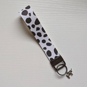 May include: A white and brown cow print key chain with a silver key ring and a silver cow charm.