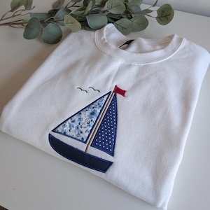 以下が含まれることがあります： 白いスウェットシャツに、前面に青と白の帆船が刺繍されています。帆船は赤い帆と船体に花柄があります。