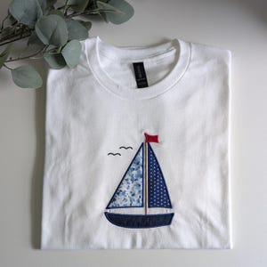 Op de afbeelding: Wit T-shirt met een geborduurd zeilbootontwerp. De zeilboot heeft een blauw bloemenprint zeil, een blauw gestippeld zeil en een rode vlag. Twee kleine zwarte vogels bevinden zich boven de zeilboot. Het T-shirt is netjes opgevouwen.