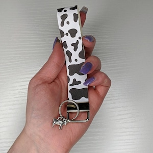 May include: A black and white cow print key ring with a silver cow charm. The key ring is attached to a silver key ring.
