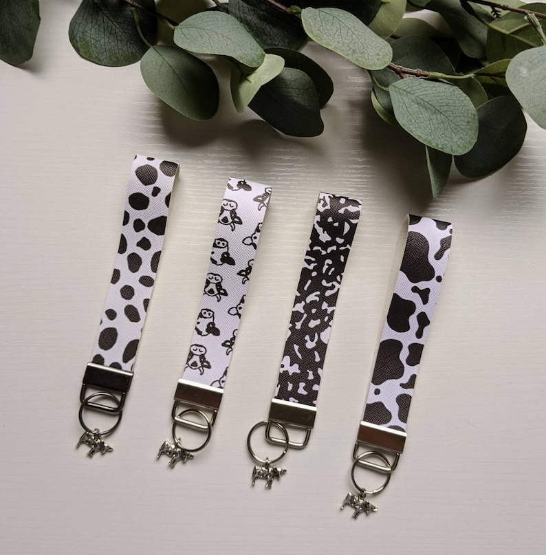 May include: Four black and white patterned keychains with cow-themed charms. The keychains feature different patterns, including cow spots, cow print, and a repeating cow illustration.