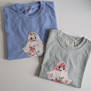 May include: Two pastel-coloured t-shirts, one light blue and one sage green, each featuring a floral ghost applique. The ghost design is made of white fabric with pink, red, and green floral patterns. The shirts are made of a soft material.