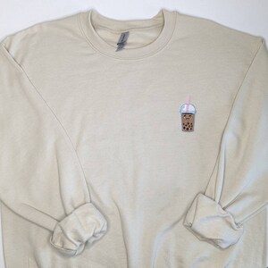 May include: A beige sweatshirt with a small embroidered image of a brown bubble tea with a pink straw and a white lid.