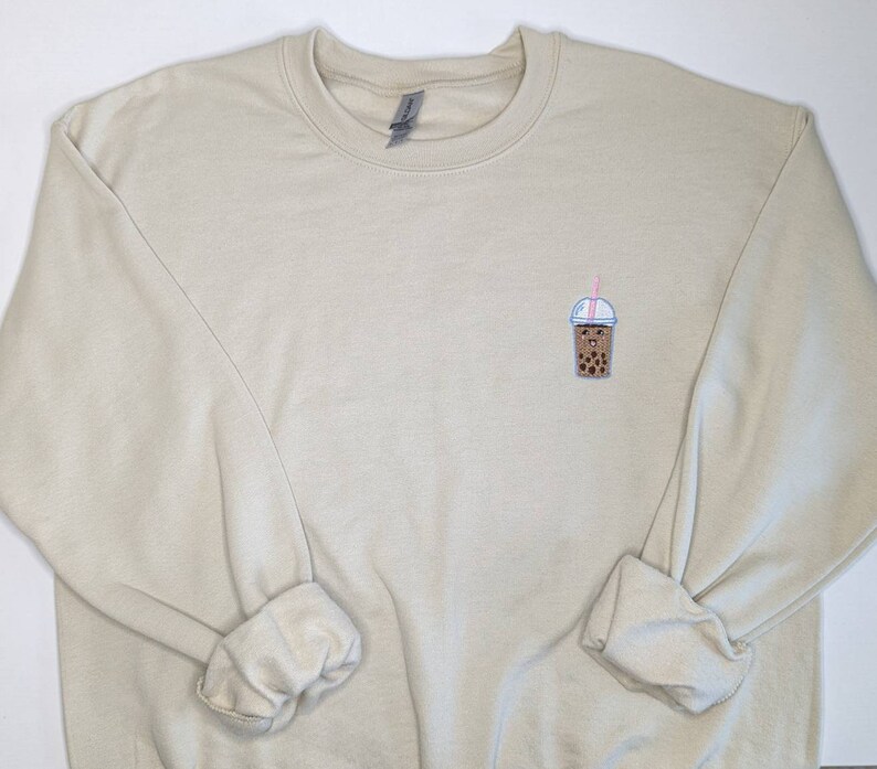 May include: A beige sweatshirt with a small embroidered image of a brown bubble tea with a pink straw and a white lid.