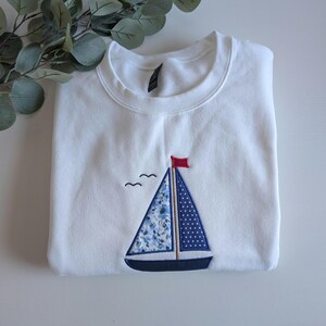 May include: White sweatshirt with a blue and white sailboat embroidered on the front. The sailboat has a red flag and a blue and white patterned sail.