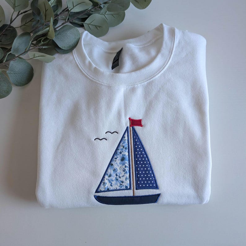 May include: White sweatshirt with a blue and white sailboat embroidered on the front. The sailboat has a red flag and a blue and white patterned sail.