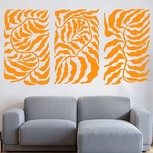 May include: Three rectangular wall art pieces with orange leaf designs against a white wall. A gray sofa and a cactus in a white pot are in front of the wall art. A floor lamp and a rug are also in the room.