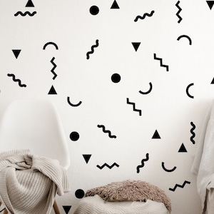 May include: A white wall decorated with black geometric shapes, including circles, triangles, and wavy lines. A white chair with a black and white throw blanket is in the foreground. A white sofa with a white throw blanket and a vase of dried flowers is in the background.