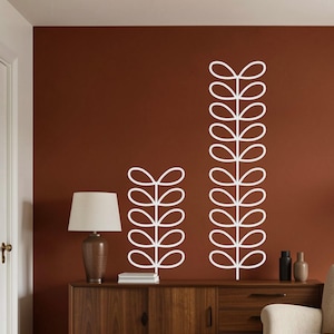 Puede incluir: Dos calcomanías de pared blancas con ramas de hojas estilizadas contra una pared marrón rojizo oscuro. Las calcomanías son de diferentes longitudes, una es más alta que la otra. Debajo de las calcomanías hay un mueble de madera con una lámpara y objetos decorativos.