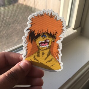 Puede incluir: Una pegatina con un personaje de dibujos animados con pelo naranja puntiagudo y una expresión angustiada. El personaje tiene la piel amarilla, la boca muy abierta y detalles azules. La pegatina se sostiene delante de una ventana.