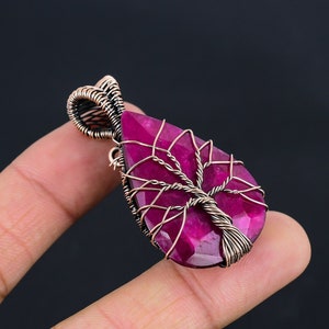 Może przedstawiać: Wisiorek w kształcie łzy z fasetowanym, głęboko różowym kamieniem szlachetnym. Kamień jest osadzony w drucie w kolorze miedzi, z motywem drzewa życia. Wisiorek ma pętlę u góry na łańcuszek. Ciemne tło.