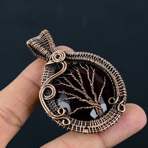 Puede incluir: Un colgante envuelto en alambre de cobre con una piedra preciosa de color rojo oscuro con un diseño de árbol. El colgante tiene un bucle en la parte superior para sujetar una cadena.