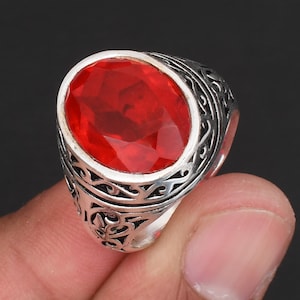 May include: A silver ring with an oval, faceted, red gemstone. The ring has an ornate, engraved design around the gemstone and on the band. The ring is held up by a person's fingers.
