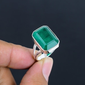 Puede incluir: Un anillo de plata con una gran piedra preciosa verde de corte esmeralda rectangular engastada en un bisel de metal pulido. La banda presenta un diseño de vástago dividido que se estrecha hacia la base, ofreciendo un aspecto equilibrado.