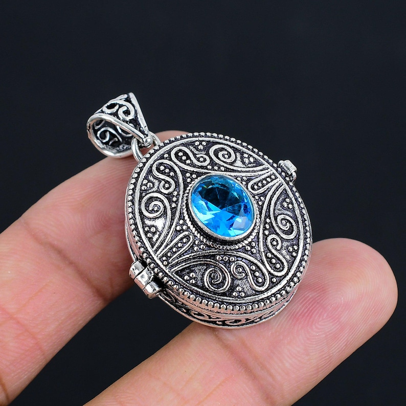 May include: An oval-shaped silver locket pendant featuring intricate embossed scrollwork patterns and a central, faceted, oval-cut bright blue gemstone. The pendant includes a decorative, ornate bail at the top for attachment to a chain.