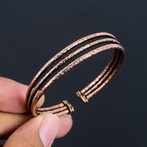 May include: A copper-toned cuff bracelet featuring three parallel bands with a textured, hammered finish. The open-ended design consists of three distinct metal wires that are joined at the terminals, creating a layered appearance with a metallic sheen.