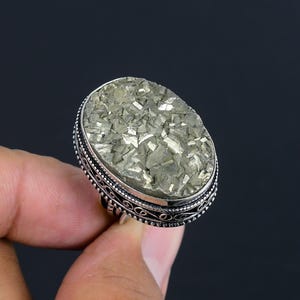 May include: An oval-shaped ring featuring a large, faceted pyrite gemstone with a metallic gold-toned finish. The stone is set in a decorative silver-colored metal band with intricate filigree scrollwork and a beaded border along the edges.