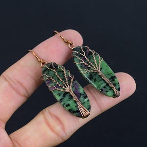 Puede incluir: Un par de pendientes de piedra verde y púrpura con un diseño de árbol de alambre de cobre. Los pendientes son de forma ovalada y tienen un bucle de alambre de cobre para colgar.